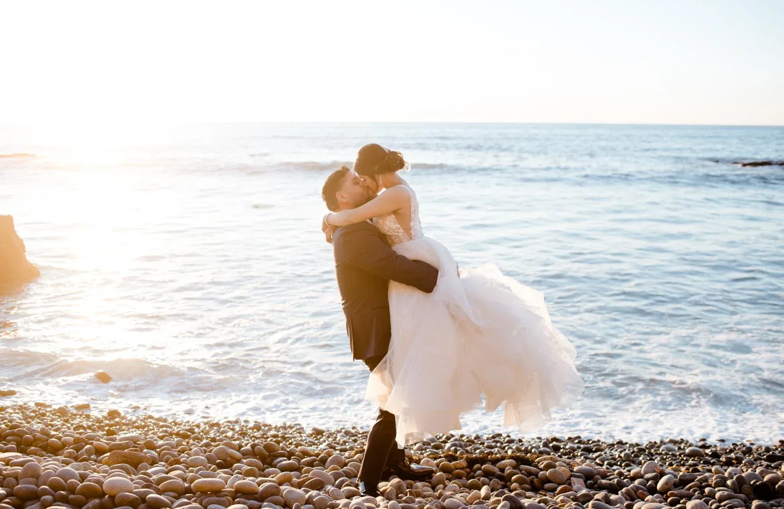 A couple embracing on a rocky beach with a white wedding gown in the breeze