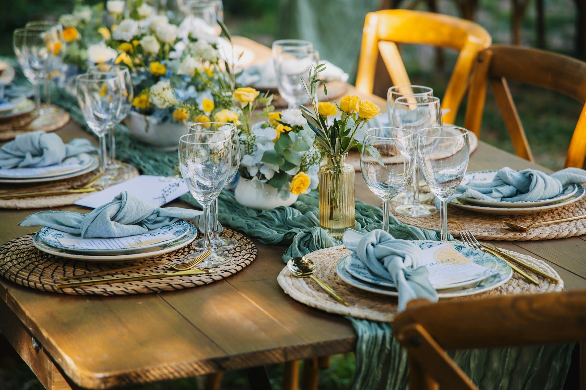 Outdoor event table layered with yellow flowers, glassware, woven place settings, and soft blue linens