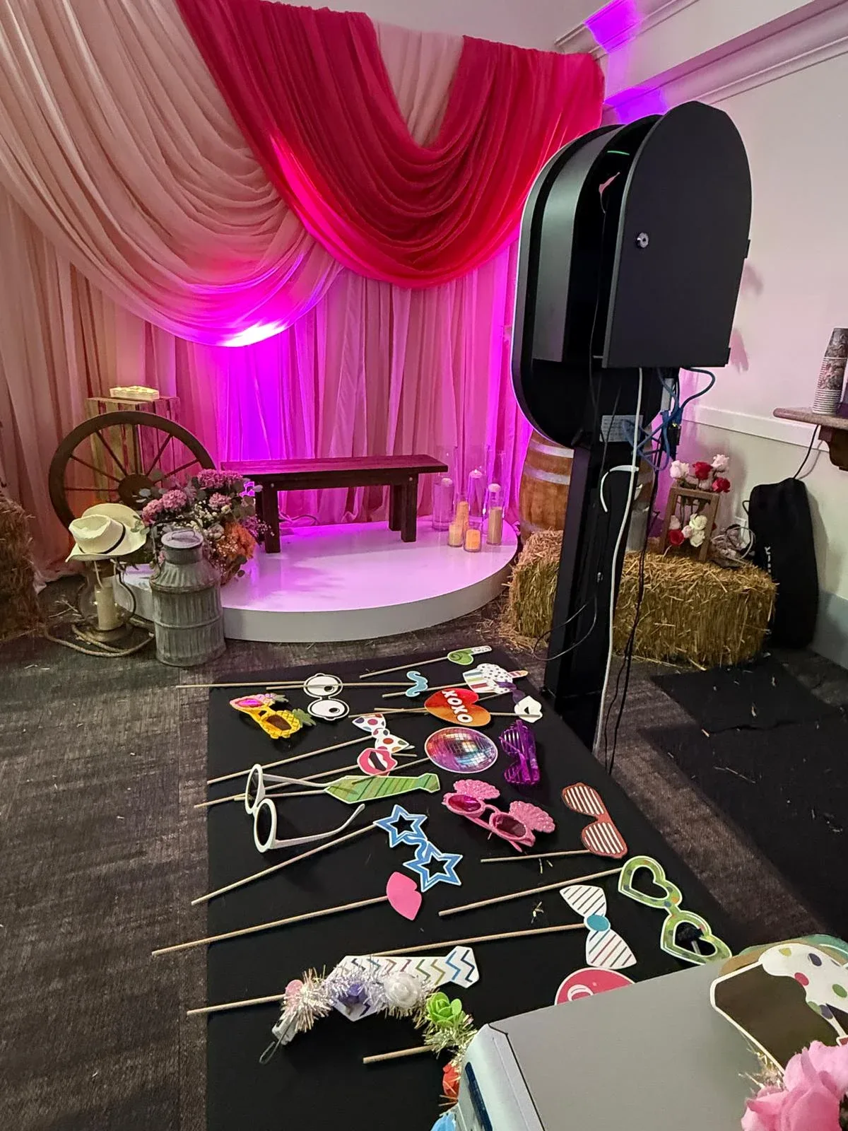 A photo booth and prop table beside a pink draped backdrop and floral arrangements
