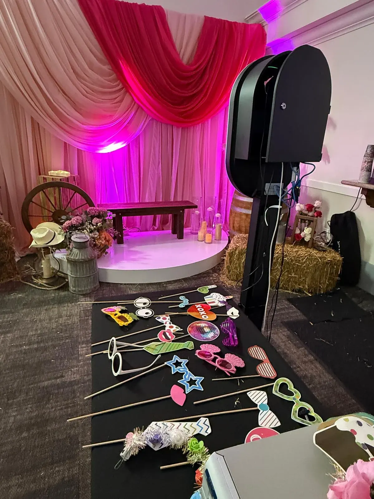 A photo booth and prop table beside a pink draped backdrop and floral arrangements