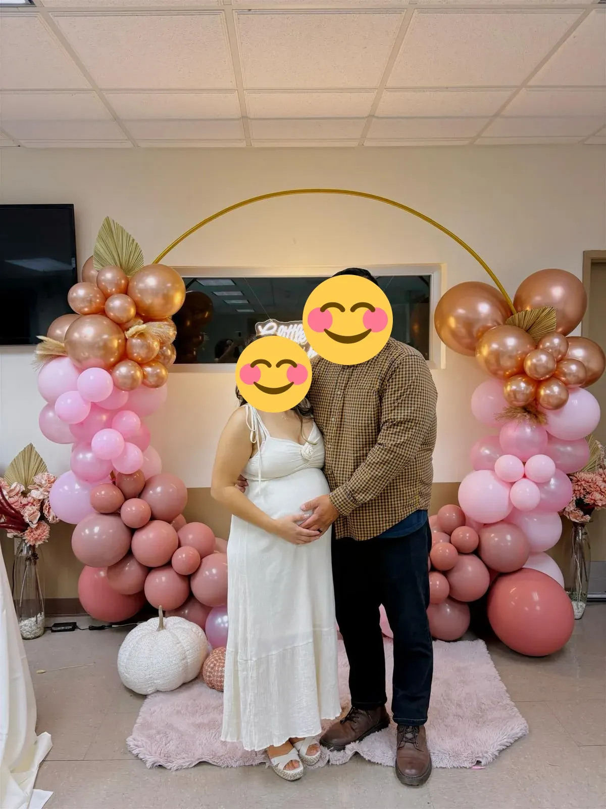 A couple at a baby shower framed by pink and copper balloons, florals, and a gold arch