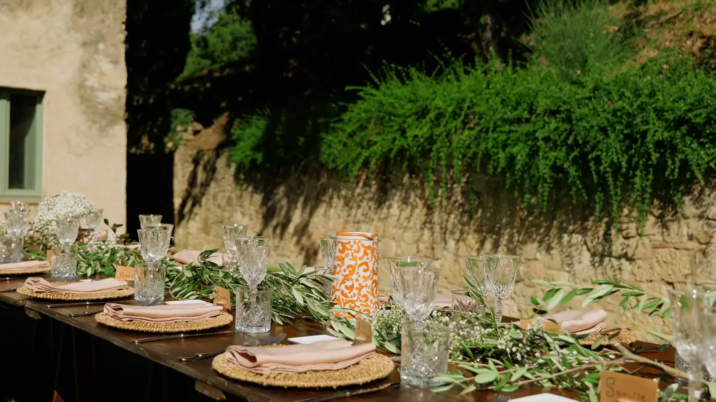 Sunlit outdoor event table arranged with greenery, flowers, glassware, and woven place settings